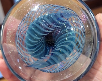 Könnte beinhalten: Ein durchsichtiger Glas-Briefbeschwerer mit einem blauen und weißen Wirbelmuster und schimmerndem Glitzer im Inneren.