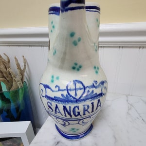 May include: White ceramic pitcher with blue and green painted details. The pitcher has two spouts and the word "SANGRIA" is painted on the front.