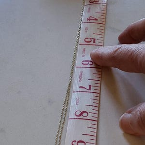 May include: A silver chain necklace with a small, round, clear gemstone pendant. The necklace is measured against a ruler, showing it is approximately 7 inches long.