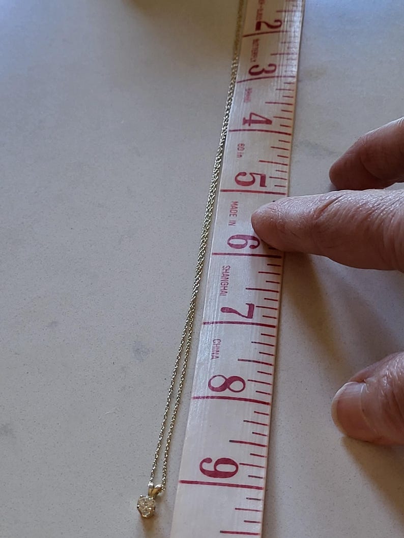 May include: A silver chain necklace with a small, round, clear gemstone pendant. The necklace is measured against a ruler, showing it is approximately 7 inches long.