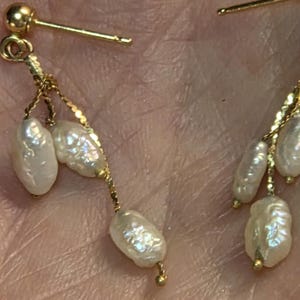 May include: Close-up of gold earrings with dangling, irregularly shaped white pearls. The earrings have a post-and-stud design with delicate gold chains connecting the pearls. The pearls have a lustrous, off-white color.
