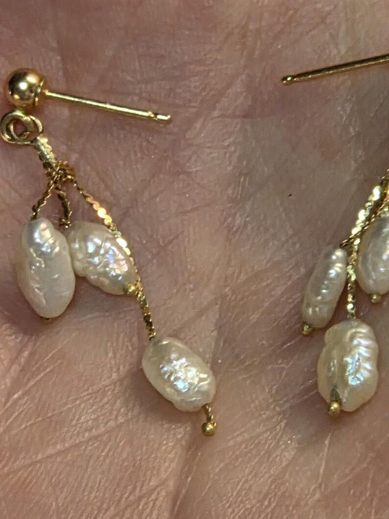 May include: Close-up of gold earrings with dangling, irregularly shaped white pearls. The earrings have a post-and-stud design with delicate gold chains connecting the pearls. The pearls have a lustrous, off-white color.