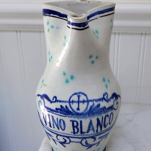 May include: White ceramic pitcher with blue and turquoise accents. The pitcher has a spout and a handle. It is decorated with a blue and white design that says "Vino Blanco" and a cross.