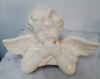May include: A white ceramic angel figurine with wings, holding its hands together in prayer. The angel has a cherubic face and is in a sitting position.