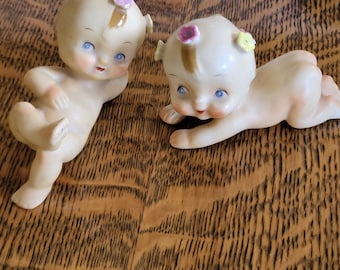 May include: Two porcelain figurines of babies with painted flower accents on their heads. The babies are nude and have blue eyes. One baby is lying on its stomach with its arms outstretched, while the other baby is sitting up with its legs crossed.