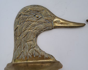 May include: A brass duck bookend with a detailed, textured design. The duck's head is turned to the side, and its beak is open. The bookend has a flat base.