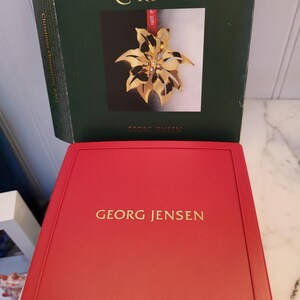 May include: A red box with the words "GEORG JENSEN" in gold lettering. The box contains a gold Christmas ornament in the shape of a poinsettia with a red ribbon. The ornament is in a green box with the words "GOLDEN CHRISTMAS" and "GEORG JENSEN" on it.