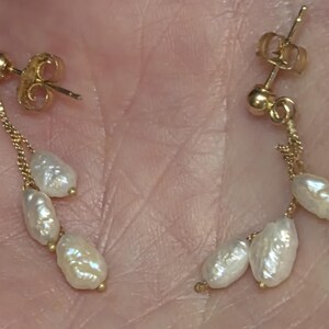May include: Gold-toned drop earrings featuring a delicate chain and three white, irregularly shaped pearls. The earrings have a post-back closure. The pearls have a lustrous, off-white color.