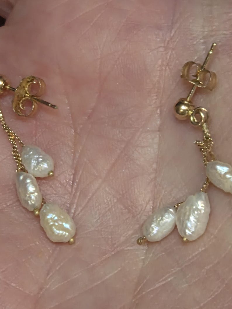 May include: Gold-toned drop earrings featuring a delicate chain and three white, irregularly shaped pearls. The earrings have a post-back closure. The pearls have a lustrous, off-white color.