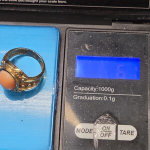 May include: A gold ring with a pink coral stone on a digital scale. The scale has a blue display and buttons labeled "MODE", "ON/OFF", and "TARE". The scale reads "Capacity: 1000g Graduation: 0.1g".
