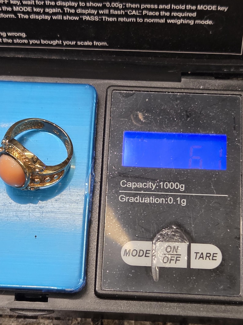 May include: A gold ring with a pink coral stone on a digital scale. The scale has a blue display and buttons labeled "MODE", "ON/OFF", and "TARE". The scale reads "Capacity: 1000g Graduation: 0.1g".