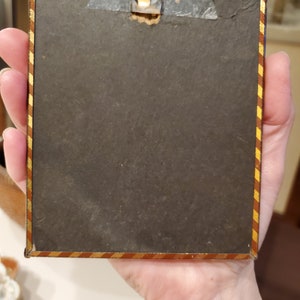 May include: A small, rectangular picture frame with a gold and red striped border. The frame has a metal hanger on the top and a black backing.