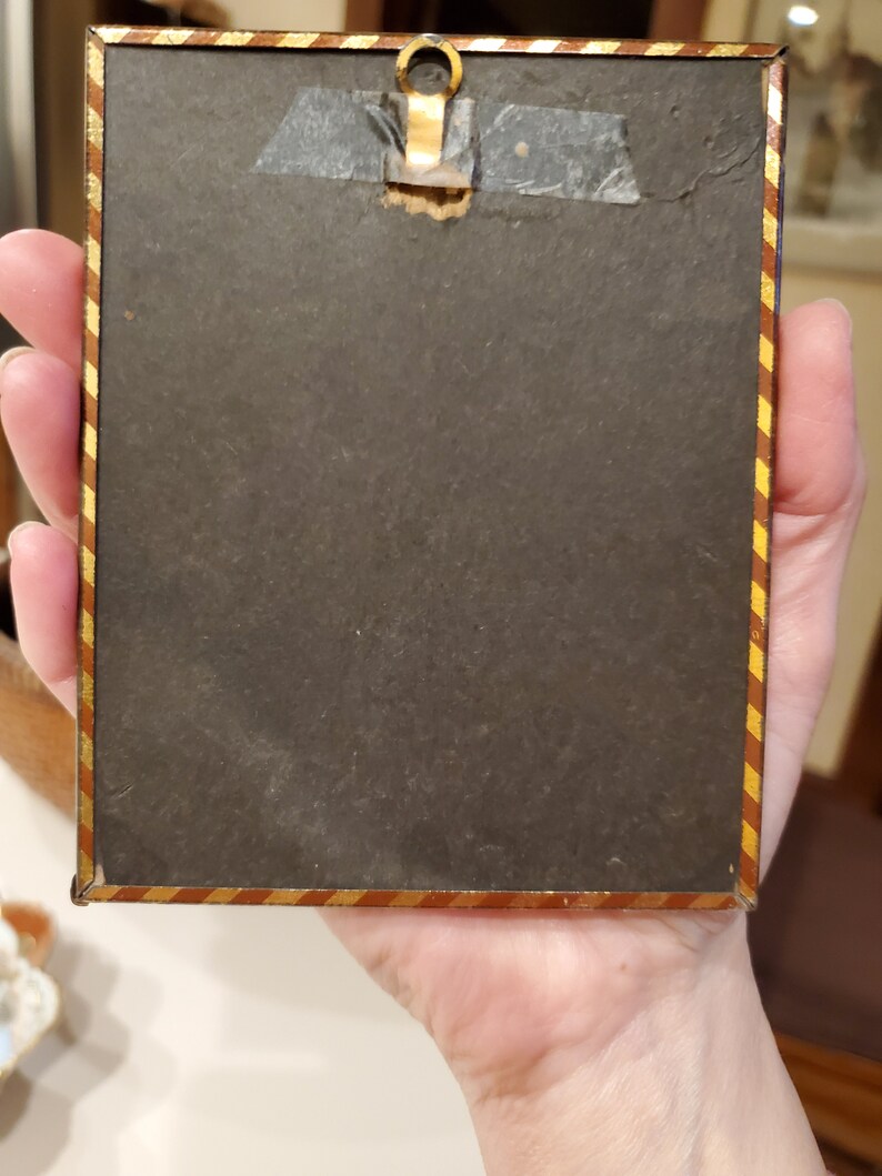 May include: A small, rectangular picture frame with a gold and red striped border. The frame has a metal hanger on the top and a black backing.