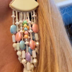 May include: A pair of long, colorful beaded earrings with a white, semi-circular top. The beads are in shades of pink, blue, green, and white.