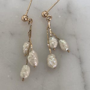 May include: A pair of gold-colored dangle earrings featuring three white, irregularly shaped pearls suspended from delicate chains. The earrings have a post-and-ball closure. The pearls have a lustrous, textured surface.