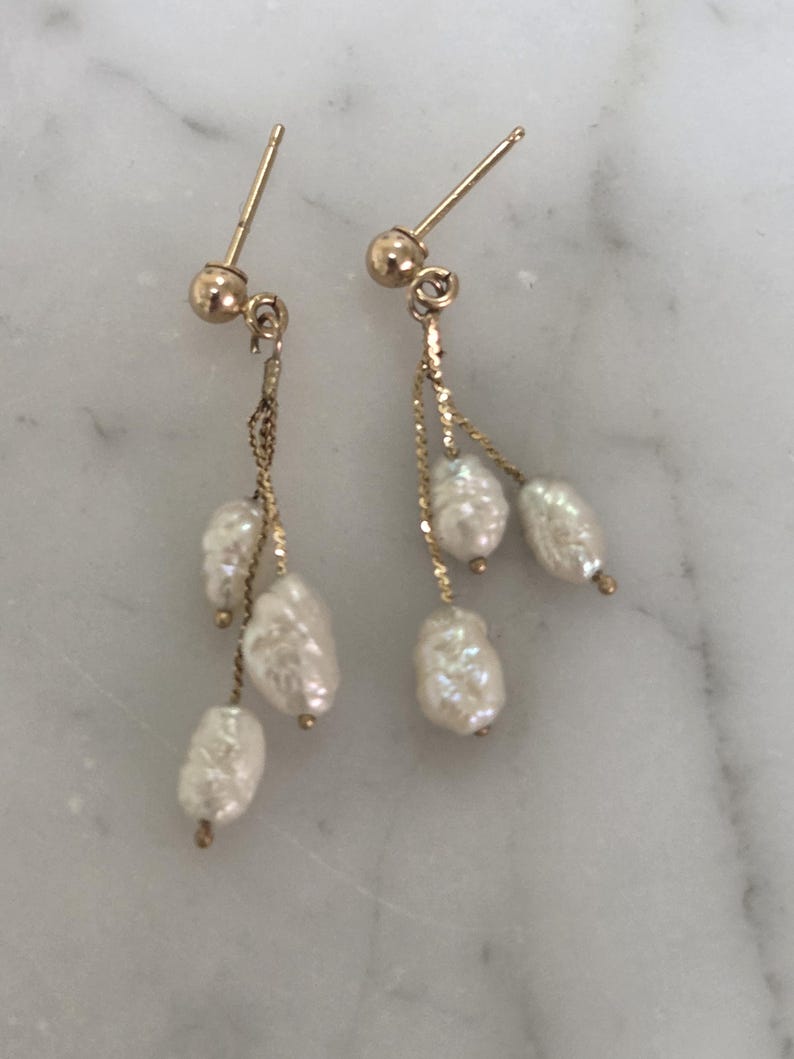 May include: A pair of gold-colored dangle earrings featuring three white, irregularly shaped pearls suspended from delicate chains. The earrings have a post-and-ball closure. The pearls have a lustrous, textured surface.
