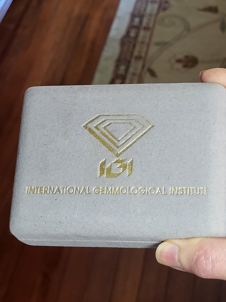 May include: A gray velvet box with the International Gemmological Institute logo in gold. The logo features a diamond shape with the letters IGI inside.