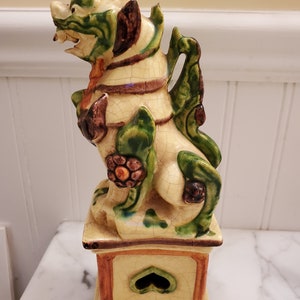 May include: A ceramic foo dog statue with green, brown, and white glaze. The foo dog is standing on a square base with a heart-shaped opening.