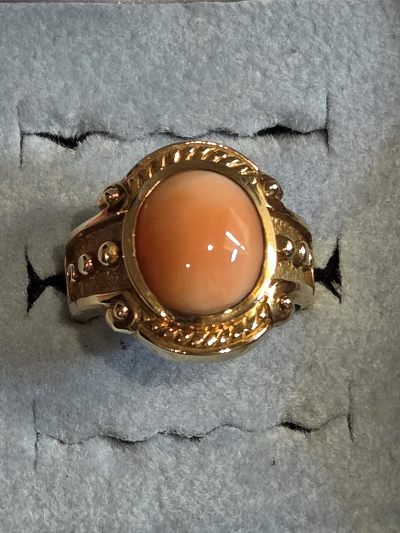 May include: A gold ring with a large oval-shaped coral stone set in a detailed bezel. The ring has a textured band with small gold beads.