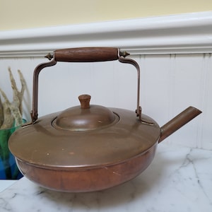 May include: A copper teapot with a wooden handle and a lid. The teapot has a round body and a spout on the side.