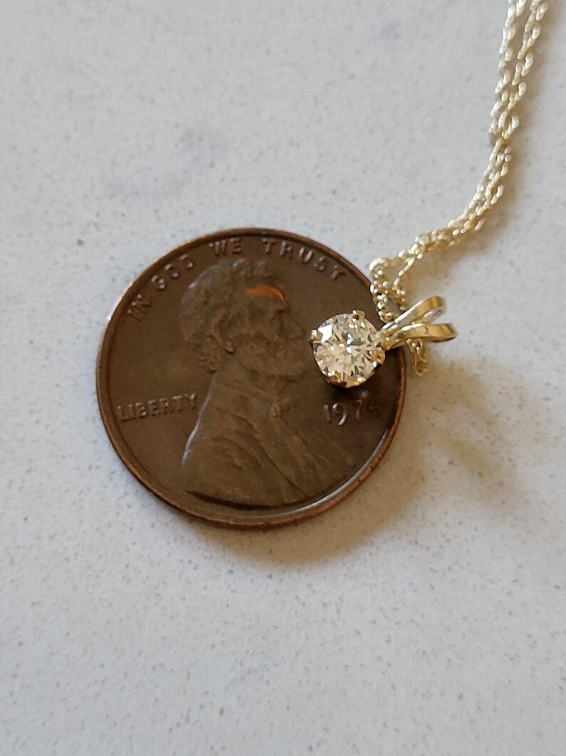 May include: A small, round, clear diamond pendant on a gold chain. The pendant is about the size of a US penny.