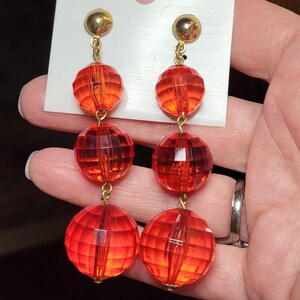 May include: A pair of long, dangling earrings with three red, faceted, round beads on each earring. The beads are connected by gold-colored metal.