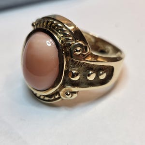 May include: A gold ring with a large, oval-shaped, light pink gemstone set in a detailed, ornate setting.