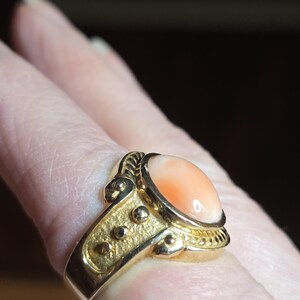 May include: A gold ring with a large oval-shaped peach coral gemstone. The ring has a detailed design with small gold balls and a rope-like pattern around the stone.