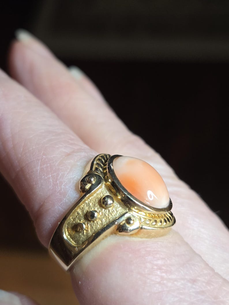 May include: A gold ring with a large oval-shaped peach coral gemstone. The ring has a detailed design with small gold balls and a rope-like pattern around the stone.