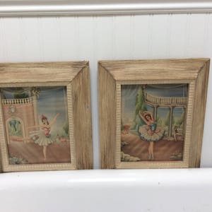 May include: Two framed pictures of ballerinas in vintage style frames. The frames are made of wood and have a light brown finish. The pictures are of ballerinas in white tutus and are set against a background of a garden.