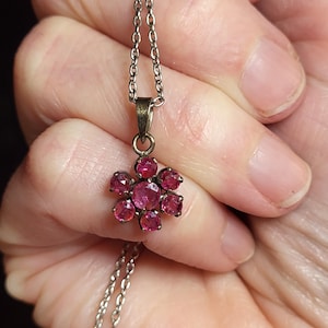 May include: A silver-toned necklace featuring a flower-shaped pendant. The pendant is adorned with multiple round, deep pink gemstones. The chain is delicate, and the pendant is held up by a small bail.