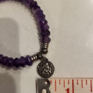 May include: A purple amethyst gemstone bracelet with a silver charm featuring a small angel figure. The bracelet is approximately 1 inch in diameter.