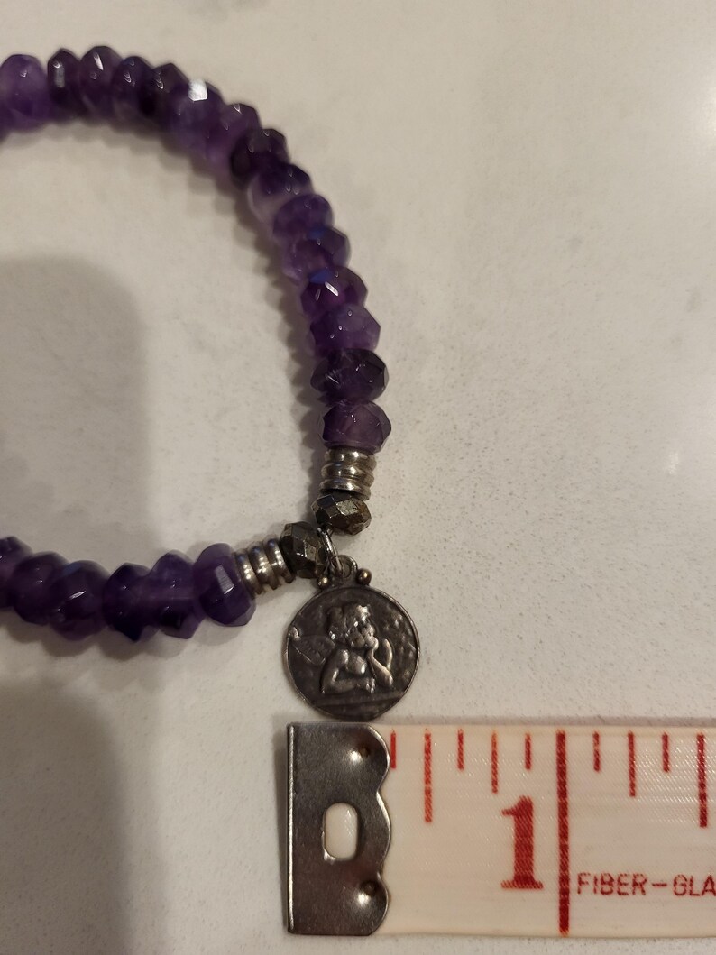 May include: A purple amethyst gemstone bracelet with a silver charm featuring a small angel figure. The bracelet is approximately 1 inch in diameter.