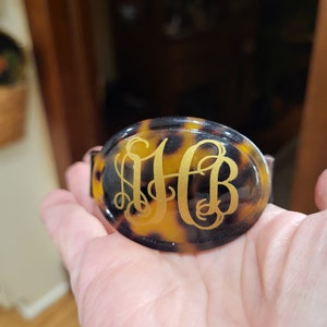 May include: A tortoiseshell-patterned hair clip with a gold monogram "HB" engraved on the front.