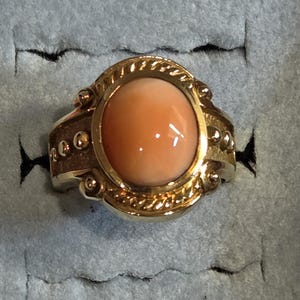 May include: A gold ring with a large oval-shaped coral stone set in a detailed bezel. The ring has a textured band with small gold beads.