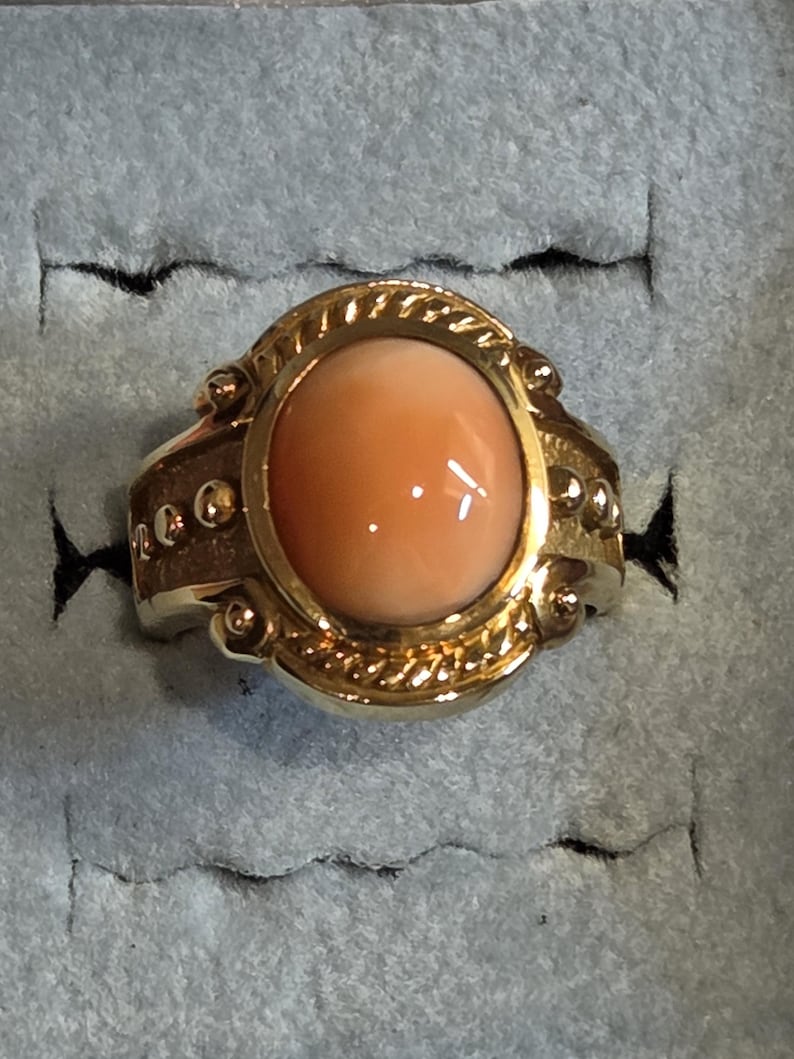 May include: A gold ring with a large oval-shaped coral stone set in a detailed bezel. The ring has a textured band with small gold beads.