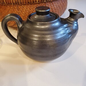 May include: A dark gray ceramic teapot with a lid and a spout. The teapot has a smooth, rounded body and a slightly textured finish.