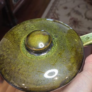 May include: Close-up of a ceramic pot lid with a dark olive green glaze. The lid features a rounded knob and a glossy finish. The glaze has variations in color, creating a textured appearance. The pot lid is a kitchenware item.