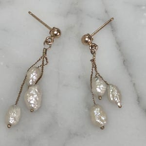 May include: A pair of gold-colored earrings with a post and a dangling chain. Each earring features three white, irregularly shaped pearls. The earrings are displayed on a white and gray marble surface.