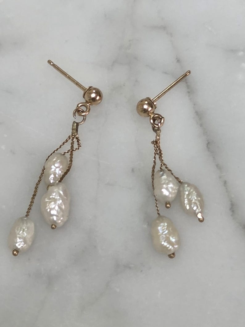 May include: A pair of gold-colored earrings with a post and a dangling chain. Each earring features three white, irregularly shaped pearls. The earrings are displayed on a white and gray marble surface.