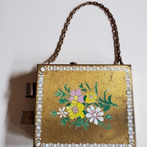 May include: A gold metal box with a floral design and a chain handle. The box has a white border with a repeating pattern of circles.
