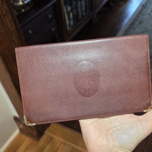 May include: A brown leather wallet with gold-colored corner accents. The wallet features a debossed logo in the center. The wallet is rectangular and appears to be closed.
