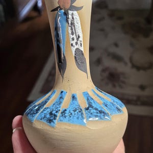 May include: A small, beige ceramic vase with a bulbous base and a narrow neck. The vase features a blue and black geometric design around the base and painted feather motifs in blue, white, and black near the top. The vase is held in a hand.