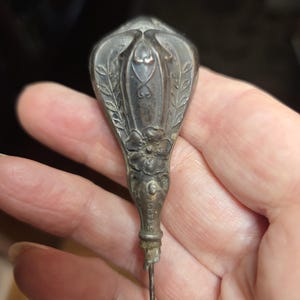 May include: Antique silver-toned hat pin with an ornate floral and leaf design. The pin's head features a detailed, symmetrical pattern with a central floral motif and delicate leaf accents. The pin is held in a hand.