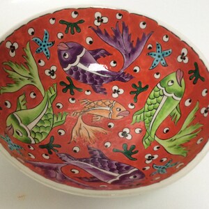 May include: A red ceramic bowl with a white rim, decorated with a colorful design of fish, flowers, and seaweed. The bowl is approximately 8 inches in diameter.