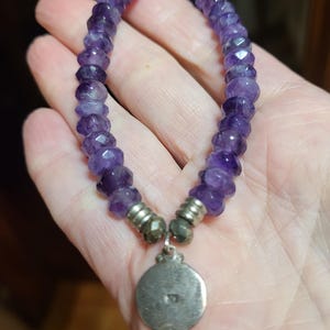 May include: A purple amethyst beaded bracelet with a silver charm. The charm is round and has a design on it.