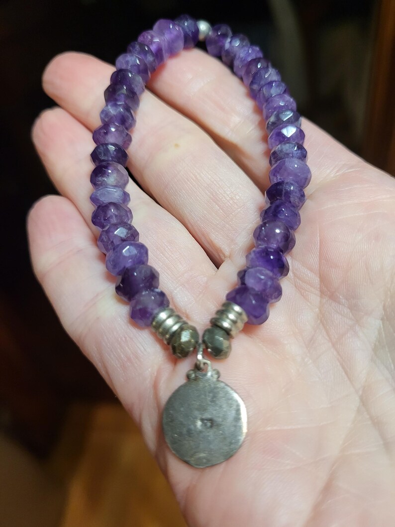 May include: A purple amethyst beaded bracelet with a silver charm. The charm is round and has a design on it.