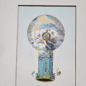 May include: An antique print of a hot air balloon, featuring a detailed illustration with figures and a landscape scene. The balloon has a blue and gold decorative base, with a yellow and white patterned top. The print is framed in a white wooden frame.