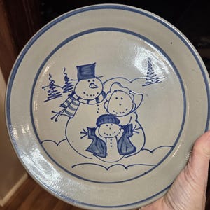 May include: A round, stoneware plate with a hand-painted design of a snowman family in blue. The plate has a light beige background with a blue border and a blue outline of the snowman family. The snowman family is wearing hats and scarves.