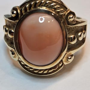 May include: A gold ring with an oval-shaped, light pink gemstone set in a rope-like design.